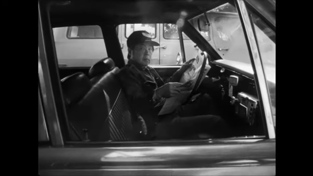 Jo (played by Wood Moy) sits in his cab, reading a newspaper. He looks out at the passenger seat.