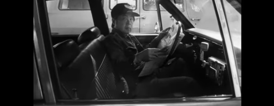 Jo (played by Wood Moy) sits in his cab, reading a newspaper. He looks out at the passenger seat.