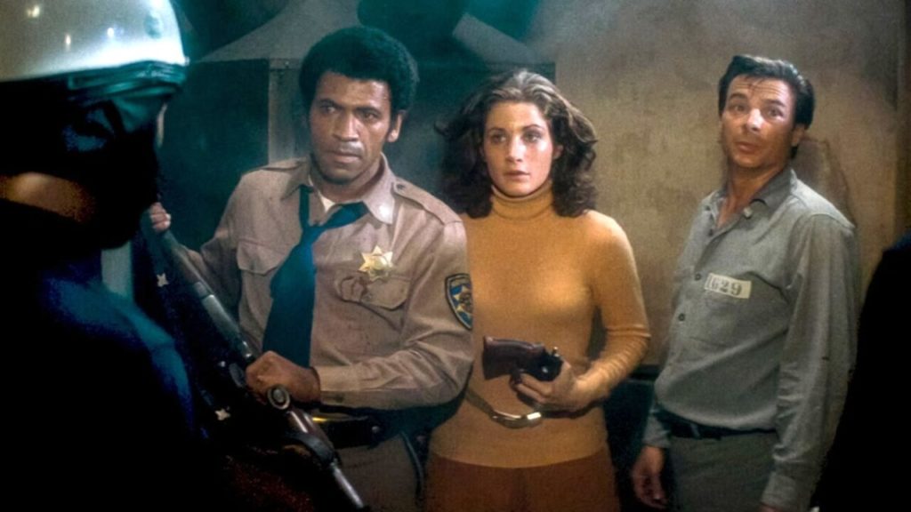 Three people -- a man with a rifle, a woman with a pistol, and a man wearing a prisoner's uniform -- stand in a smoky room, staring at a uniformed police officer. 