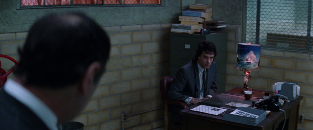 Tom Hanks as Joe, seated at a small desk with a garish lamp in a gray, drab room, in the film "Joe Versus the Volcano."