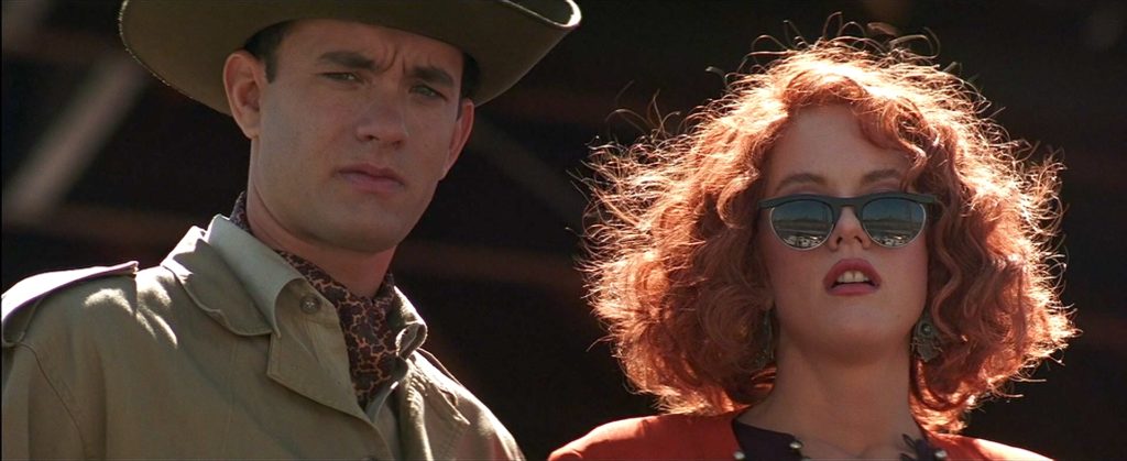 Tom Hanks wearing a hat standing next to Meg Ryan with red hair wearing sunglasses, both of them looking toward the camera in the film "Joe Versus the Volcano."