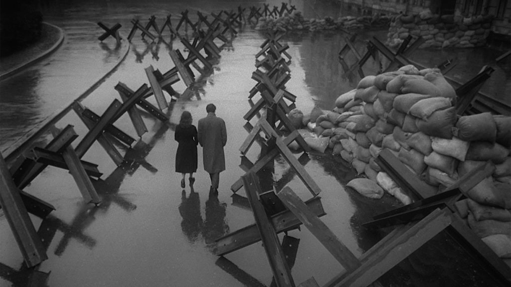 A man and a woman walk down a wet surface with barricades on either side of them in the film "The Cranes Are Flying."