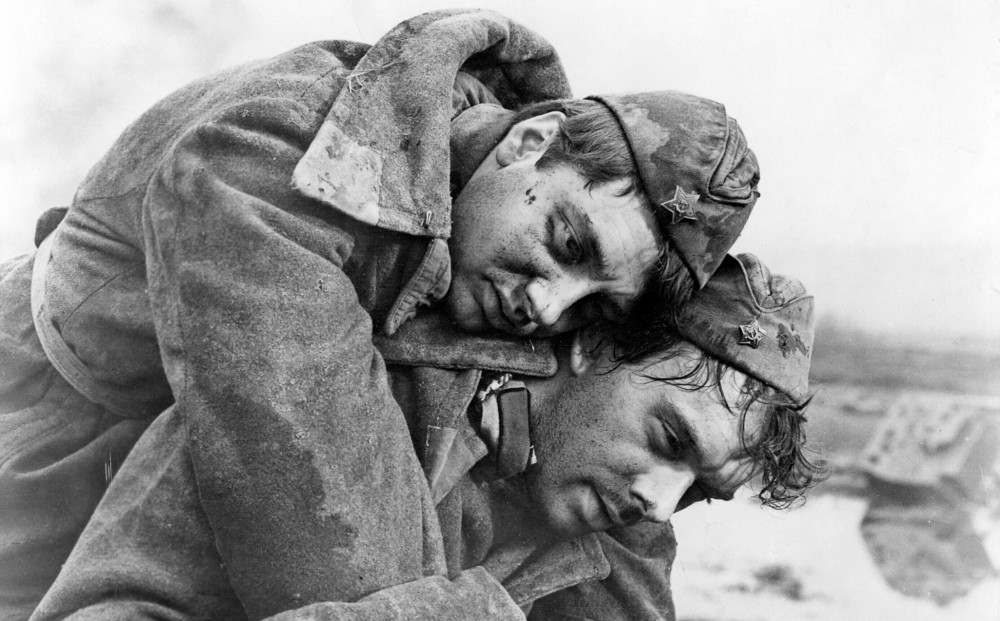 A soldier carries another soldier on his back, both of them bloodied and dirty, in the film "The Cranes Are Flying."