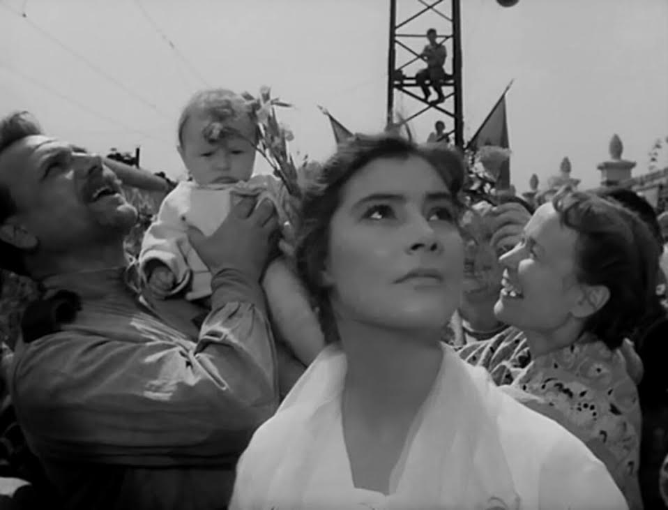 A woman looks up at the sky with an ambiguous expression as a celebration goes on behind her in the film "The Cranes Are Flying."