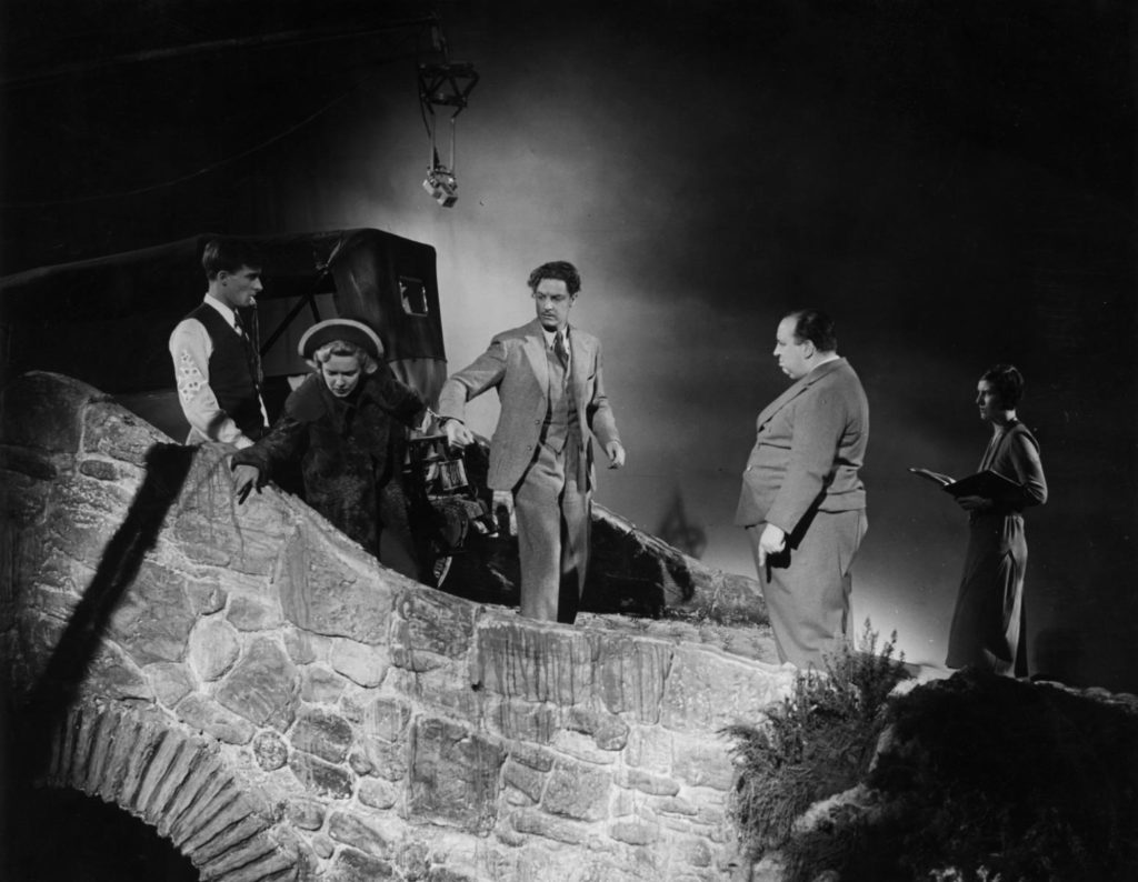 A black-and-white image of five people standing on a bridge on a movie set with a microphone visible above them. From left to right are a dark-haired white man with a whistle in his mouth; actress Madeleine Carroll, a blonde white woman; actor Robert Donat, a dark-haired, white man with a mustache; director Alfred Hitchcock, a balding white man; and a dark-haired woman holding a large book.