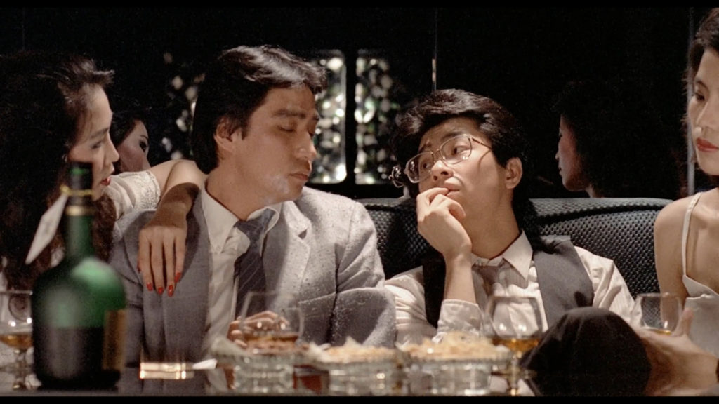 Two clean-shaven men in business suits are flanked by a pair of black-haired Asian women at a restaurant table. The man on the right wears a pair of glasses and looks at the other man with a weary expression.