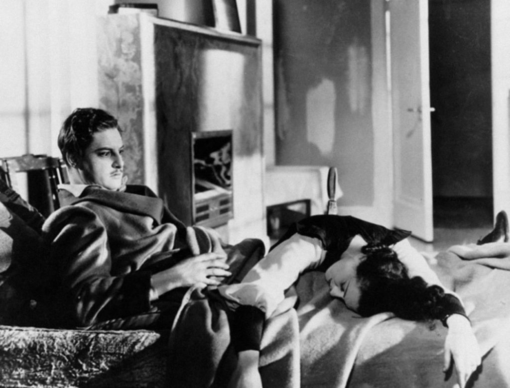 A black-and-white image of Annabella Smith (actress Lucie Manheim), a dark-haired white woman, lying across the lap of Richard Hannay (actor Robert Donat), a dark-haired white man with a mustache. Smith has the handle of a knife sticking out of her back, and Hannay looks surprised.