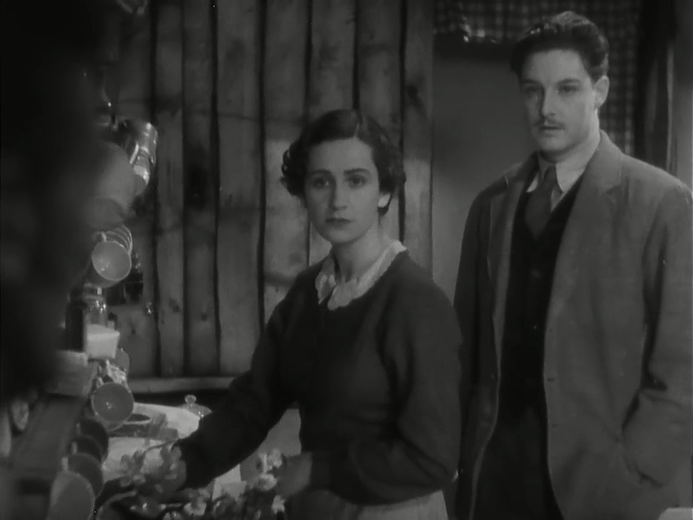 A black-and-white image of Margaret (actress Peggy Ashcroft), a dark-haired white woman, standing in front of Richard Hannay (actor Robert Donat), a dark-haired white man with a mustache. Both Margaret and Hannay are looking directly ahead.