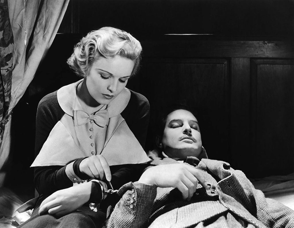 A black and white image of Pamela (actress Madeleine Carroll), a blonde white woman, sitting on a bed next to Richard Hannay (actor Robert Donat), a dark-haired white man with a mustache. Pamela tries to open the handcuff that ties her hand to Hannay’s while he is asleep.