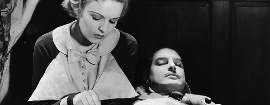 A black and white image of Pamela (actress Madeleine Carroll), a blonde white woman, sitting on a bed next to Richard Hannay (actor Robert Donat), a dark-haired white man with a mustache. Pamela tries to open the handcuff that ties her hand to Hannay’s while he is asleep.