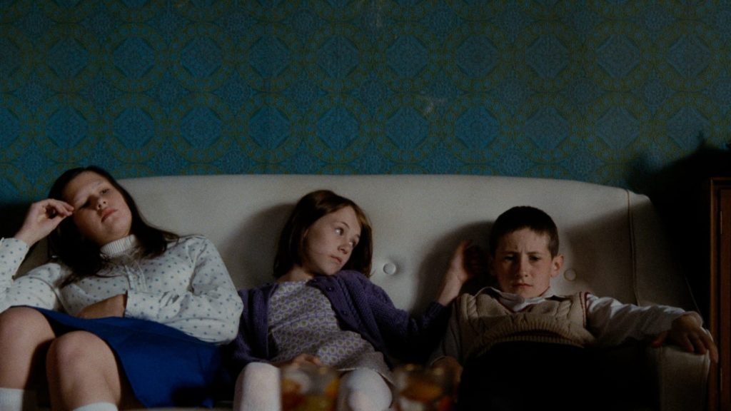 Living in a large family, Jamie (right) struggles to stand out and earn the affection of his family. Here, he sits on a couch next to his sisters, with diamond-shaped wallpaper behind them. 