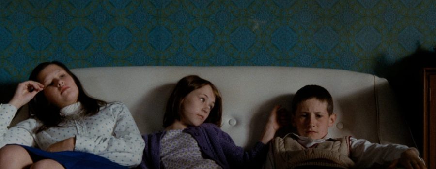 Three children sit on a couch in a dark room with diamond-patterned wallpaper in the film "Ratcatcher."
