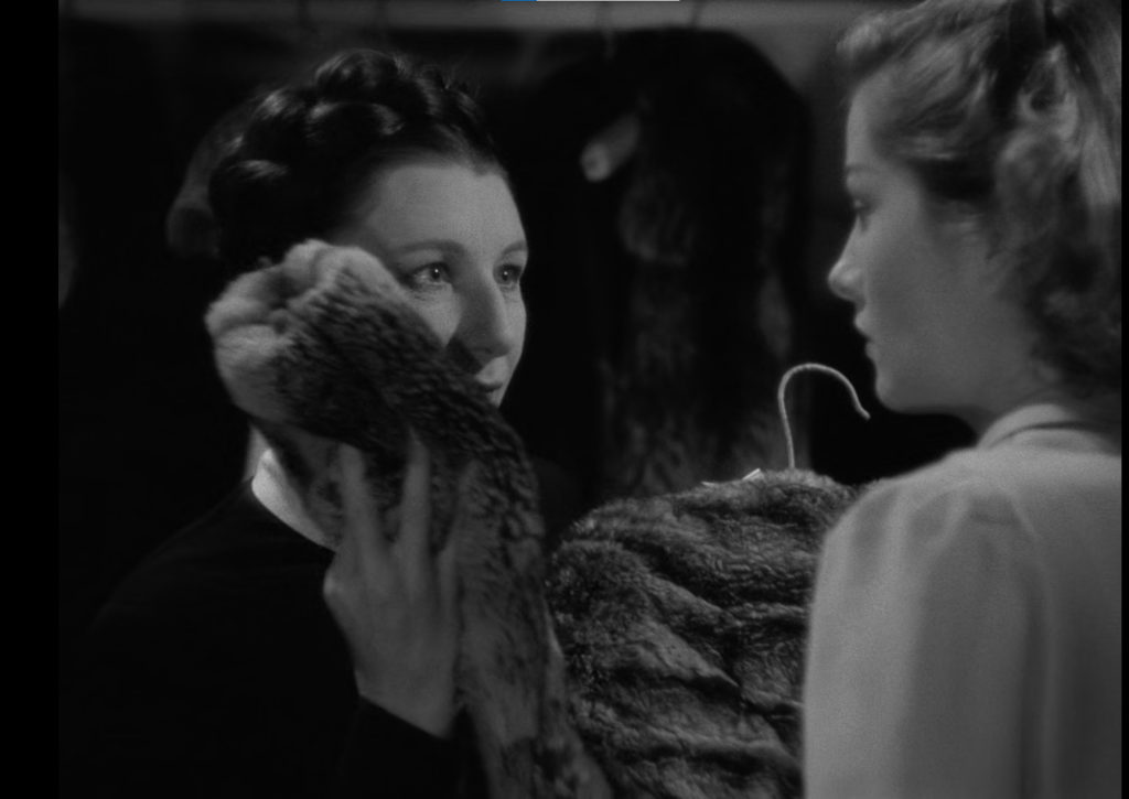Judith Anderson holds the sleeve of a fur coat to her cheek while looking at Joan Fontaine in the film Rebecca.
