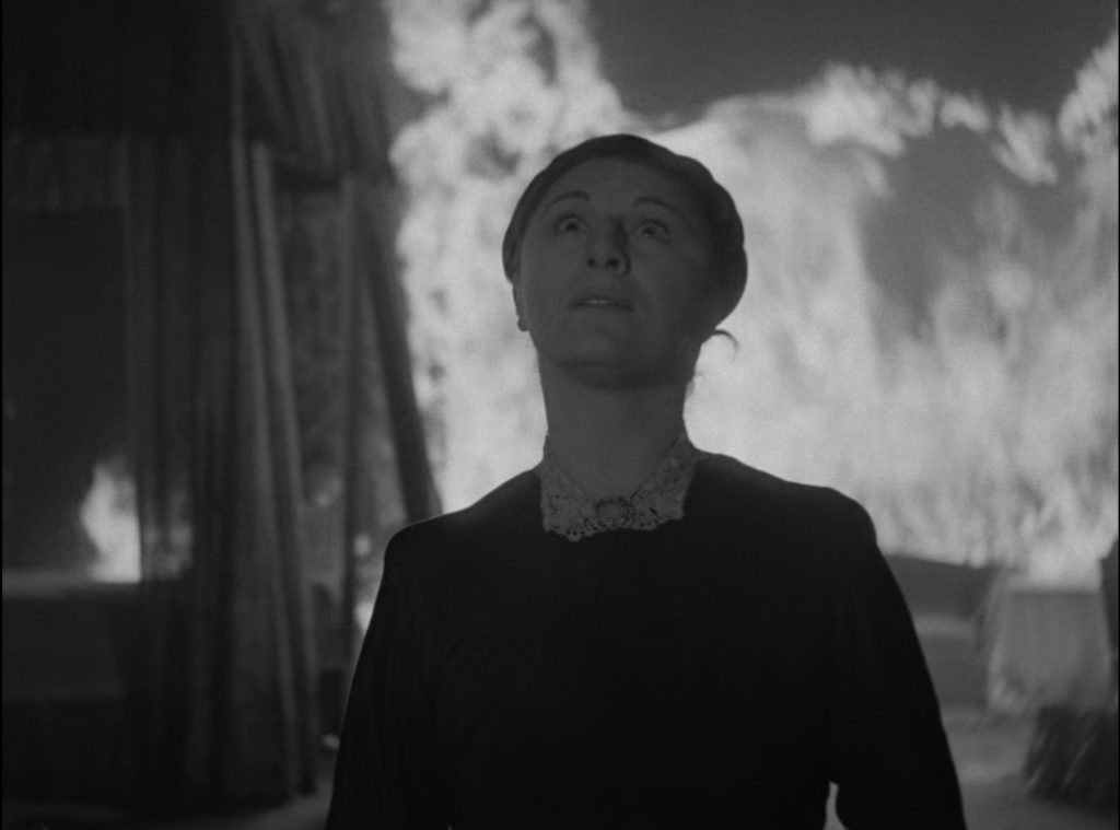 Judith Anderson looks to the heavens with an expression of fear and awe while a bedroom burns around her in the film Rebecca.