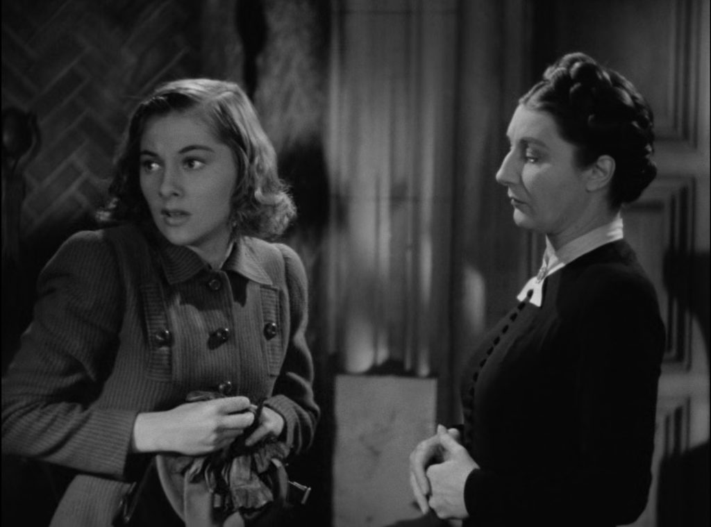 Joan Fontaine looks over her shoulder with anxiety while Judith Anderson looks at her with disapproval in the film Rebecca.