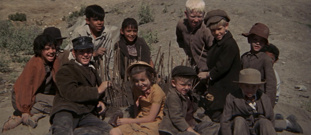 A group of kids in old-fashioned clothes sits on the sand and dirt, looking toward the camera. 