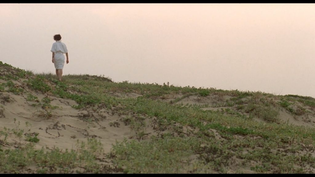 A woman with black hair and wearing a white dress is walking up toward the crest of a sand dune covered in grass.