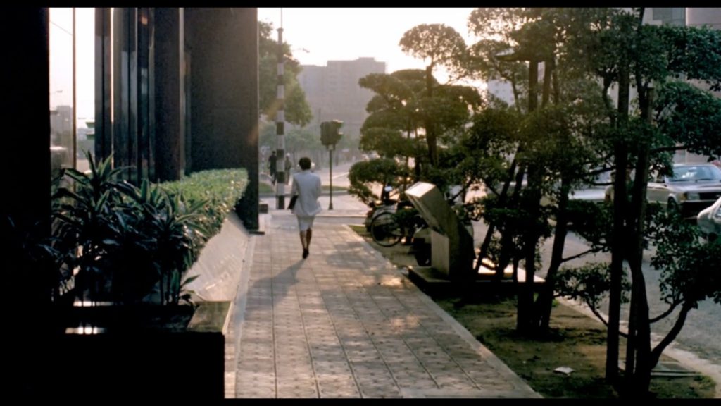 A woman in a white business suit is walking down a street in Taipei in the middle of the afternoon. Her back is turned toward us and the sunlight casts the sidewalk in a soft glow.