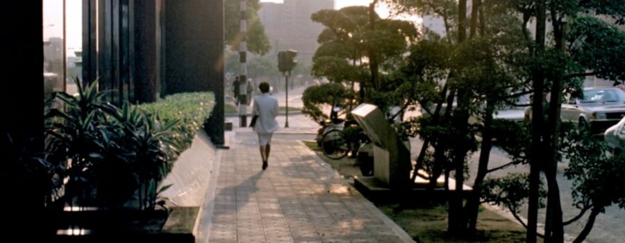A woman in a white business suit is walking down a street in Taipei in the middle of the afternoon. Her back is turned toward us and the sunlight casts the sidewalk in a soft glow.