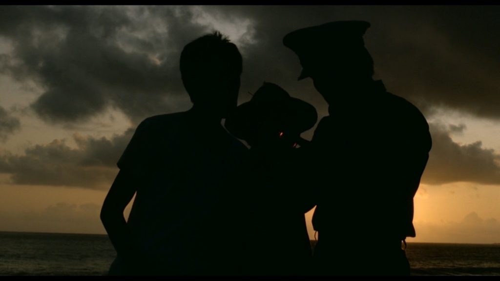 Three men are standing in silhouette against a red sunrise. One of the men is holding an object which has two red lights on the label.
