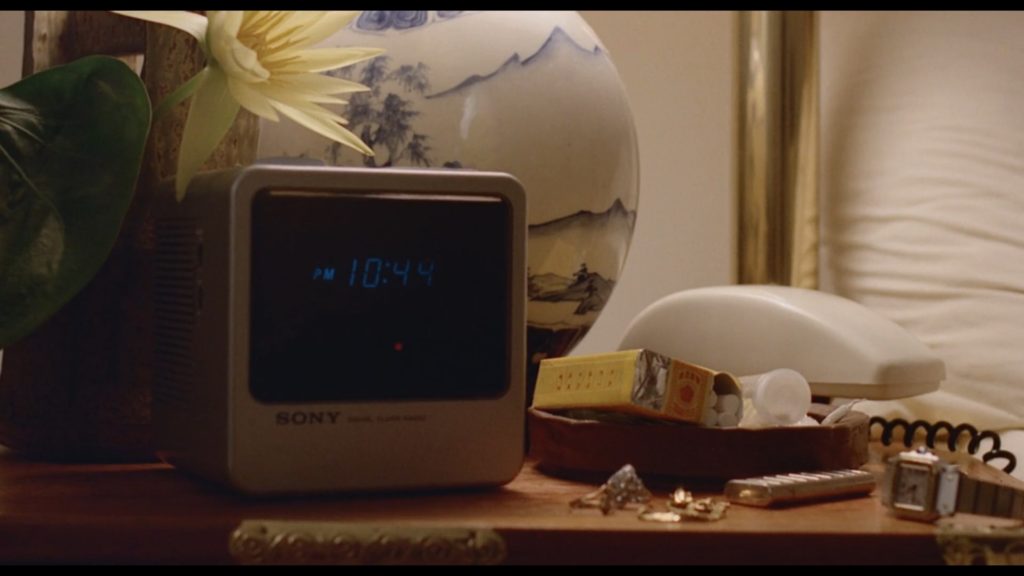 A digital clock reading 10:45 is placed beneath a yellow flower and a lamp made of blue and white china. To the right are a small watch, and an empty, crumpled-up box of cigarettes.