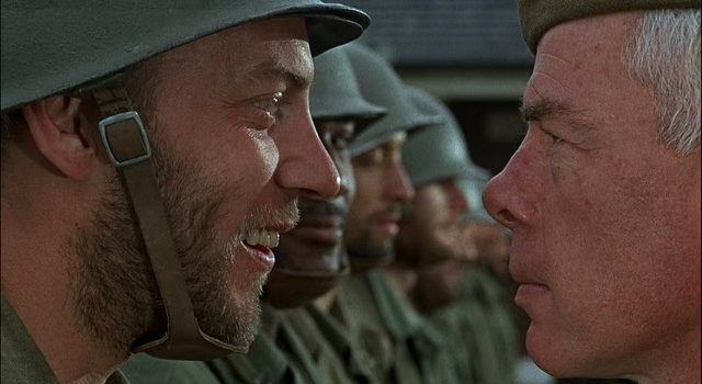 A close-up as Donald Sutherland smiles at Lee Marvin, who has a grim expression, in the film The Dirty Dozen.