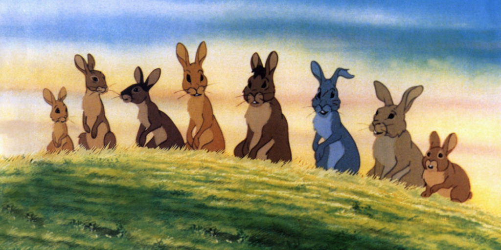 Eight rabbits sit on top of a grassy hill, a sunlit yet cloudy sky behind them, in "Watership Down."