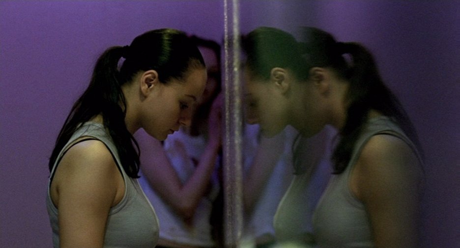 Samantha Morton as Morvern Callar with her head against the wall of a public bathroom. She is wearing a teal tank top and the walls behind her are purple. There is a woman past Morvern partially blocked from view by Morvern, out of focus.