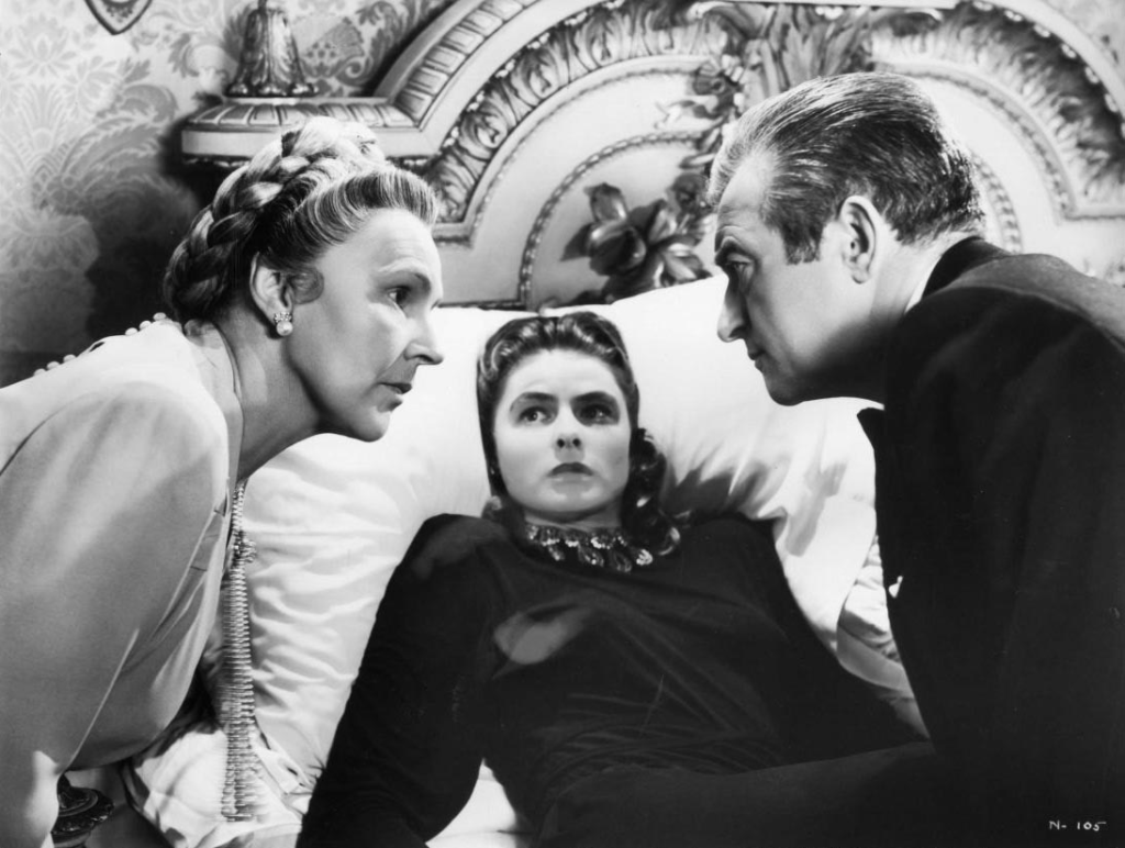 Alicia Huberman, played by Ingrid Bergman, lies on a bed looking frightened as Alexander Sebastian, played by Claude Rains, and his mother, played by Leopoldine Konstantin, look at each other with concern on either side of Alicia.