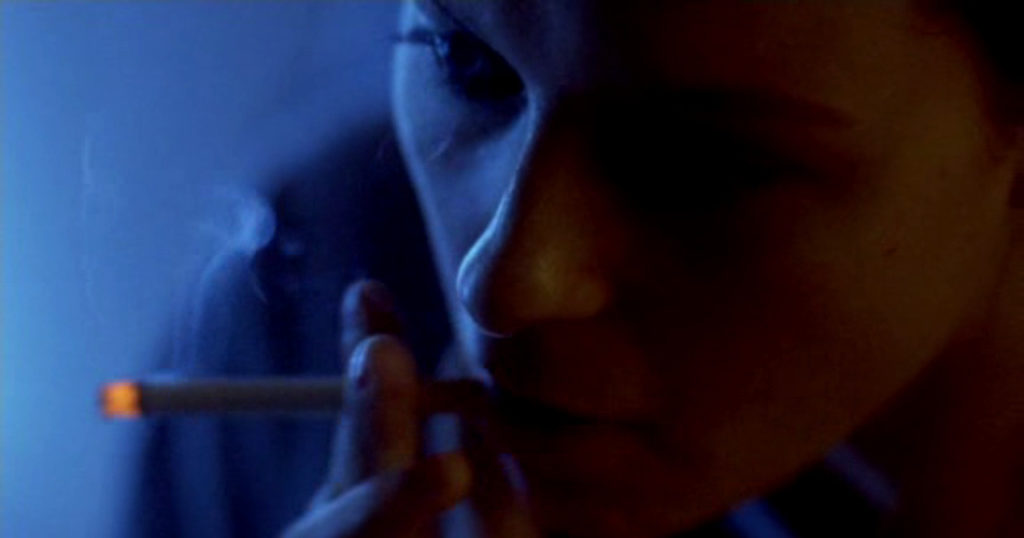 Samantha Morton as Morvern Callar smoking a cigarette. The camera is angled above her head and looks down on her, focusing on her red nail polish on her fingers. At the top of the frame, her bright blue eyes stare off ahead.