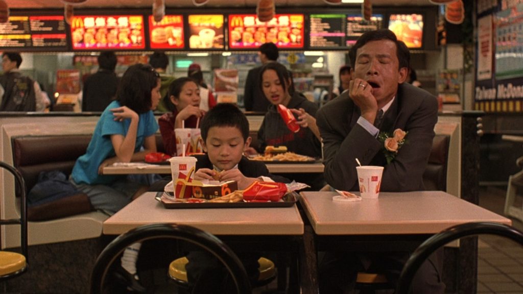 Father and eight-year-old are formally dressed in suits sitting at a table in McDonald’s in Taiwan, as seen in the film Yi Yi. The son is eating a chicken nugget from a tray of food in front of him; the father is sitting next to the son on his left and is yawning with closed eyes. 
