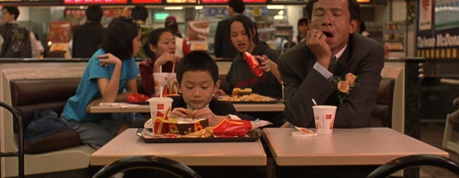 A father and his eight-year-old son are formally dressed in suits, sitting at a table in a McDonald’s in Taiwan, as seen in the film "Yi Yi." The son is eating a chicken nugget from a tray of food in front of him; the father is sitting next to the son on his left and is yawning with closed eyes.