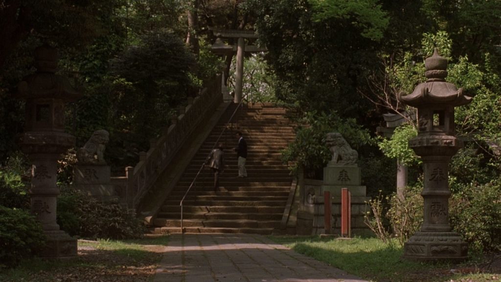 A couple climbs stairs in a leafy green park. The stairs are covered in the shadows of trees. The couple is located at a distance from the camera. 