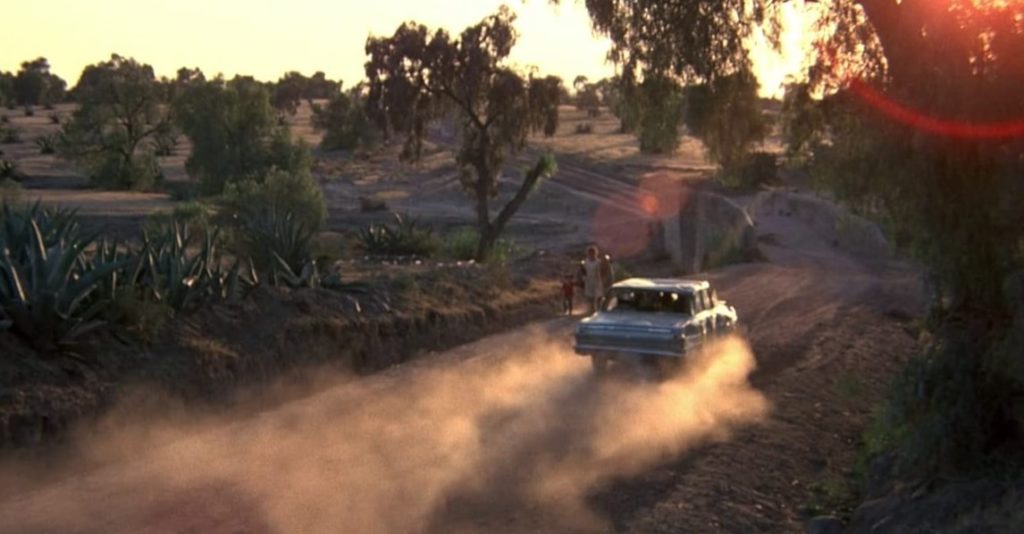 A car speeds down a dusty road at sunset.