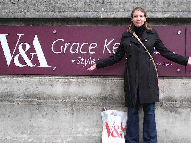 A woman wearing a black trench coat and blue jeans standing in front of a sign that says "V&A Grace Kelly Style."