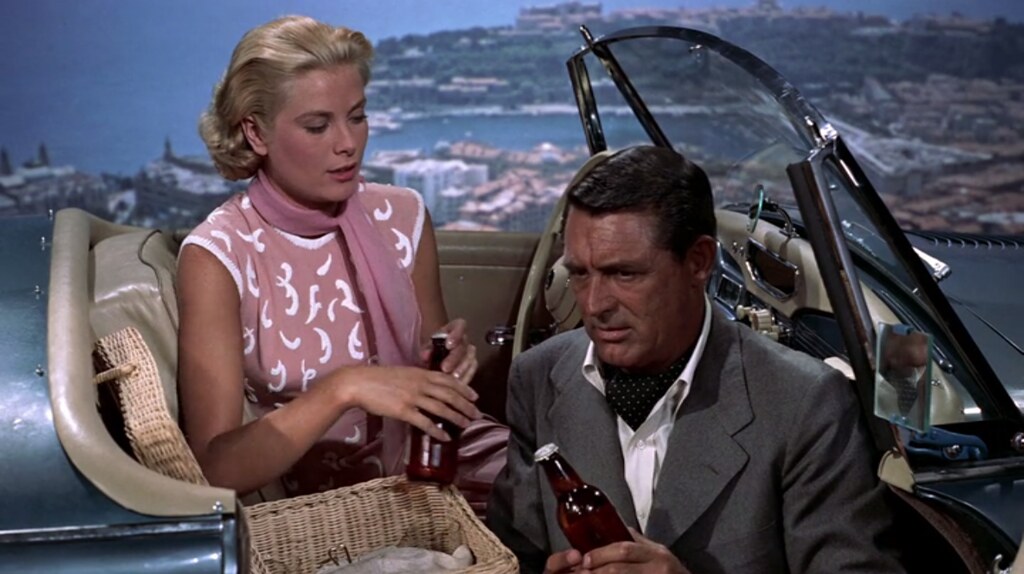 Grace Kelly and Cary Grant in a convertible with a picnic basket. Grace Kelly is wearing a peach dress with white trim and a peach scarf and Cary Grant is wearing a gray sport jacket with a white shirt and a polka-dot cravat.