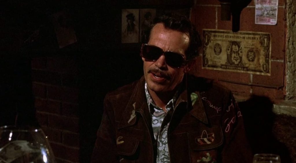 Bennie sits in a dimly lit bar, wearing sunglasses and a patterned jacket.
