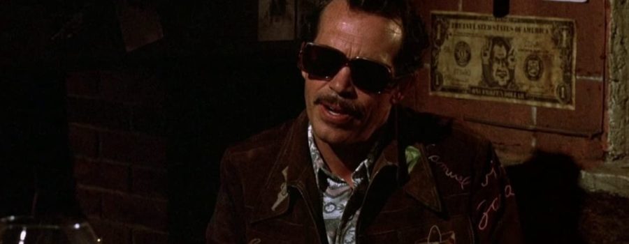 Bennie sits in a dimly lit bar, wearing sunglasses and a patterned jacket.