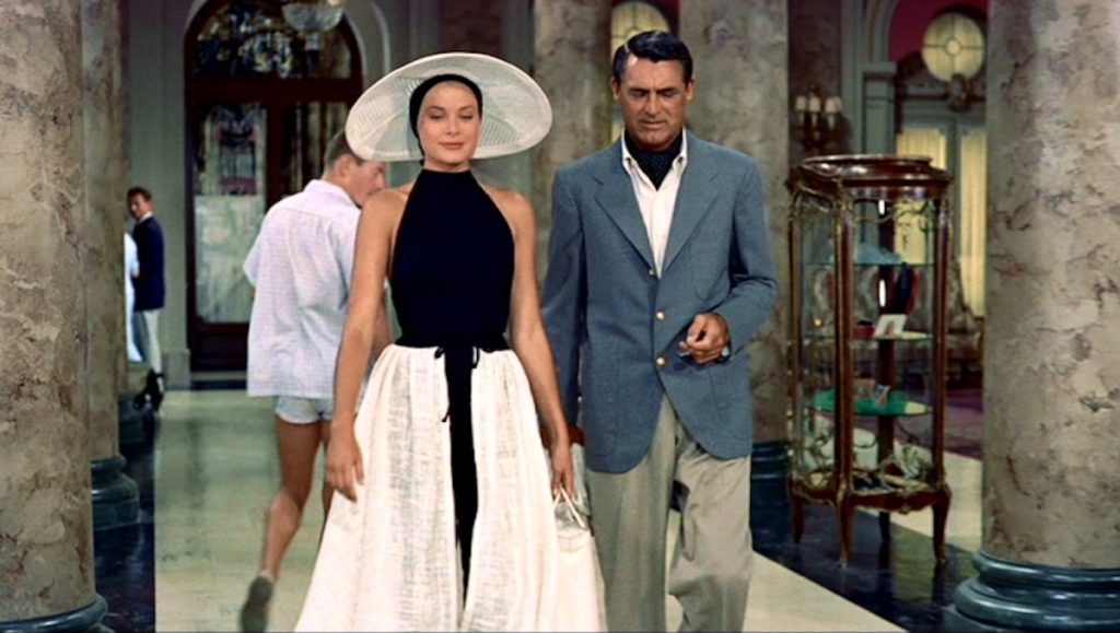 Grace Kelly and Cary Grant standing in a hotel lobby. Grace Kelly is wearing a black halter top with a white wrap skirt and a black and white sun hat. Cary Grant is wearing a gray sport jacket with tan trousers and a white shirt.