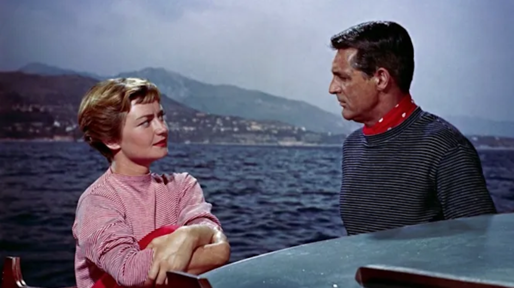 Brigitte Auber and Cary Grant on a boat with the ocean and mountains in the background. Brigitte Auber is wearing a red and white striped t-shirt and Cary Grant is wearing a navy blue and white striped t-shirt with a red and white polka-dot cravat.
