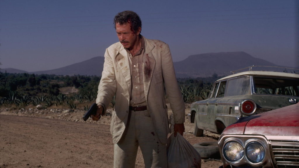 Bennie, white male in dirty beige suit, pointing a pistol with right hand, holding bag with Head of Alfredo Garcia in left hand.