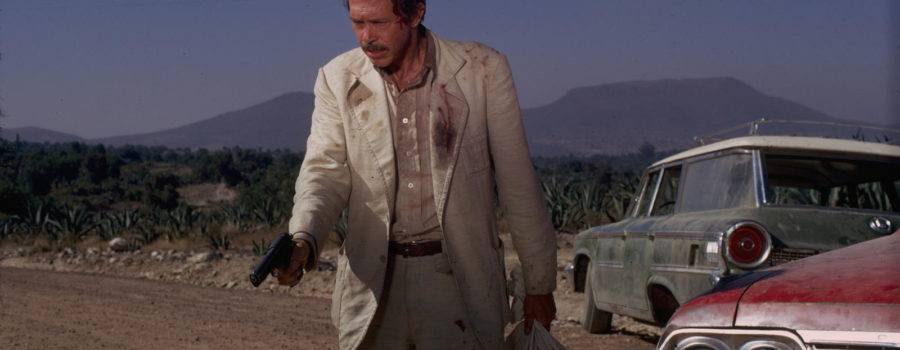 Bennie, white male in dirty beige suit, pointing a pistol with right hand, holding bag with Head of Alfredo Garcia in left hand.