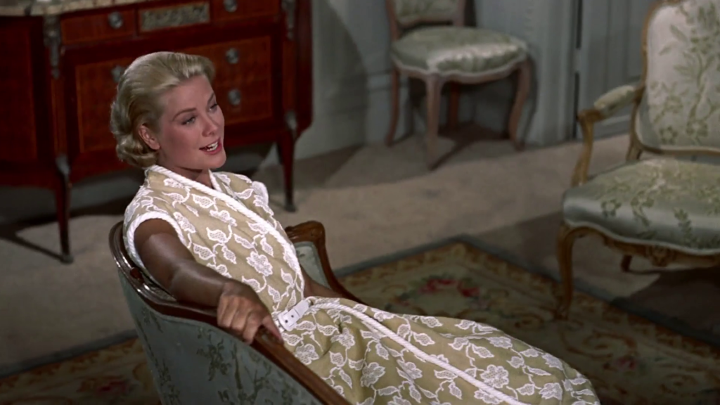 Grace Kelly lounging in a chair wearing a beige and white floral wrap dress with white trim.