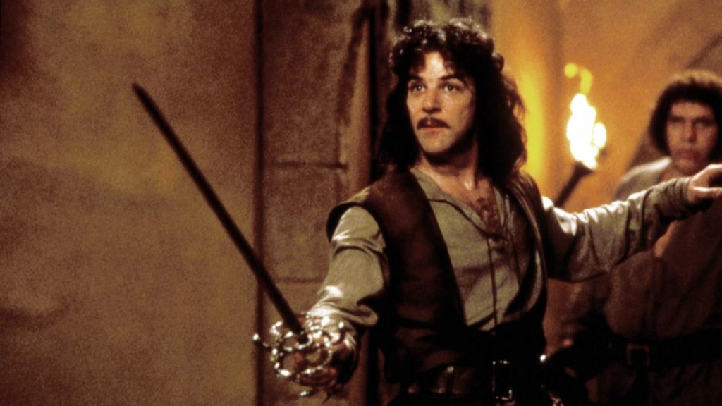 Inigo Montoya brandishing his sword, left hand raised to the side, with Fezzik out of focus in the background.