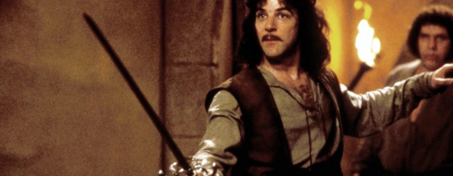 Inigo Montoya brandishing his sword, left hand raised to the side, with Fezzik out of focus in the background.