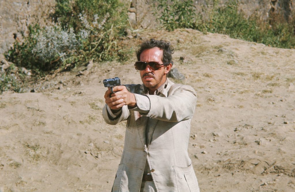 A man with a mustache and sunglasses, wearing a light-colored suit, stands in the desert, pointing a gun offscreen.