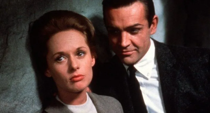 Tippi Hedren leans against a wall, looking offscreen, as Sean Connery stands behind her and looks at her in "Marnie."