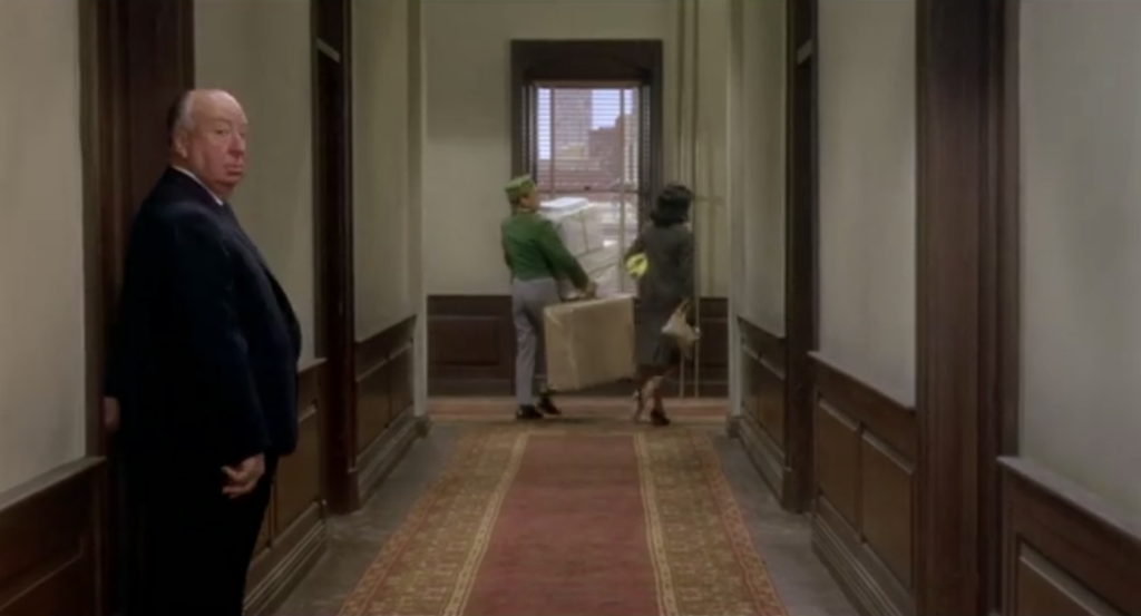 A man in a suit (Alfred Hitchcock) turns to face the audience as a woman and a bellboy in a hotel walk away from the camera down a hallway. 