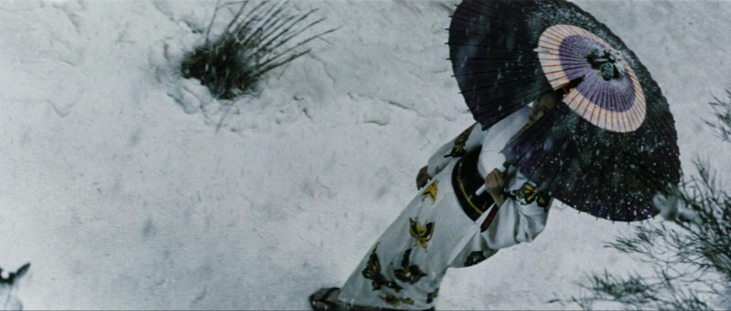 Downward-angled shot of Yuki walking through the snow in a white kimono with large yellow butterfly print, her face slightly visible through a fashionable v-cut in the purple umbrella she’s holding overhead.