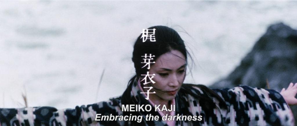 Yuki training atop jagged cliffs overlooking the ocean during the film’s title sequence, with Meiko Kaji’s name on screen. Subtitled lyrics from her song “Shura No Hana” reads, “Embracing the Darkness.”